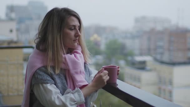 Mujer caucásica permanece en el balcón durante la lluvia con una taza de café caliente — Vídeos de Stock