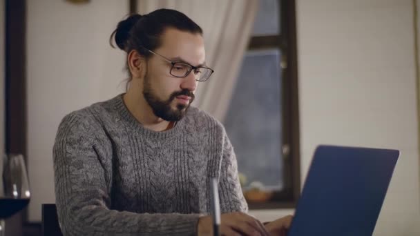 Un joven guapo con gafas trabaja en un portátil con una copa de vino. — Vídeos de Stock