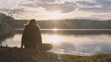 Kadın, batan güneşin ışınlarında, suyun yanındaki gölün kıyısında oturur.