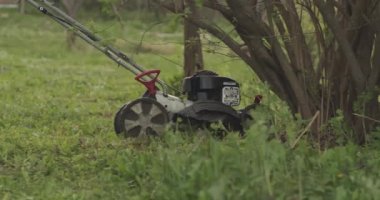 Bahçede yeşil çimenler ve elinde çim biçme makinesi olan bir adam geçiyor.