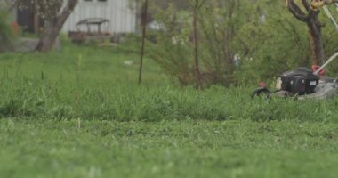 Bahçede yeşil çimenler ve elinde çim biçme makinesi olan bir adam geçiyor.