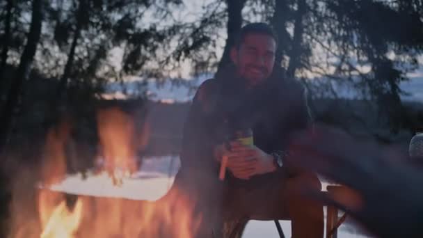 Mladý muž v bundě sedí v noci u ohně s hrnkem horkého čaje.. — Stock video
