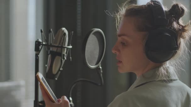 En ung flicka i monitorhörlurar framför en mikrofon spelar in en podcast — Stockvideo
