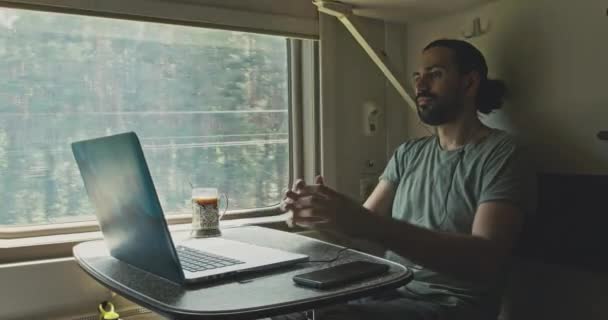 Un joven caucásico se sienta en auriculares con una computadora portátil mientras viaja en tren. — Vídeos de Stock