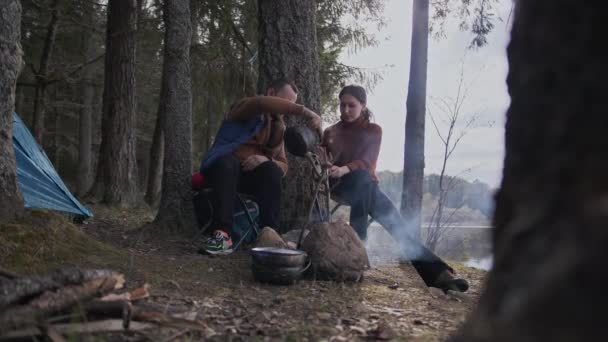 En un día de otoño, los turistas amorosos se sientan junto al fuego y beben té de tazas de hierro. — Vídeos de Stock