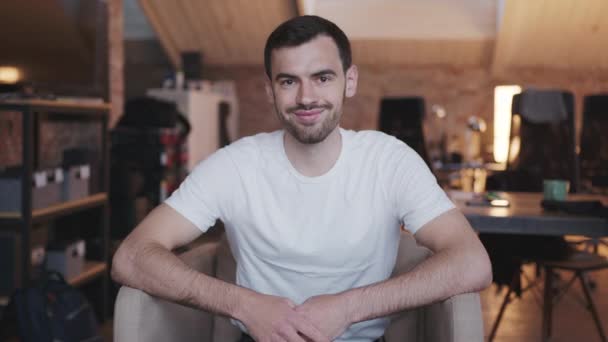 Ein junger Mann im weißen T-Shirt sitzt auf einem Stuhl, blickt in die Kamera und lächelt.. — Stockvideo