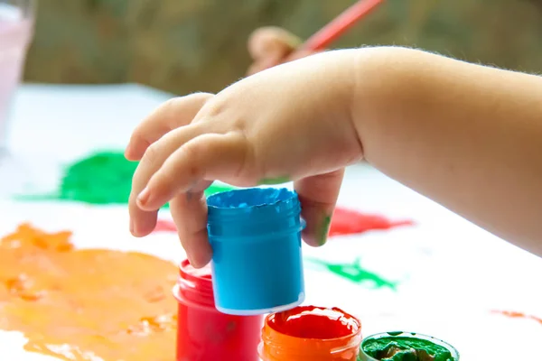 A child\'s hand holds a jar of paint about to draw with it. Smears of bright paint on white paper. The brush in the paint.