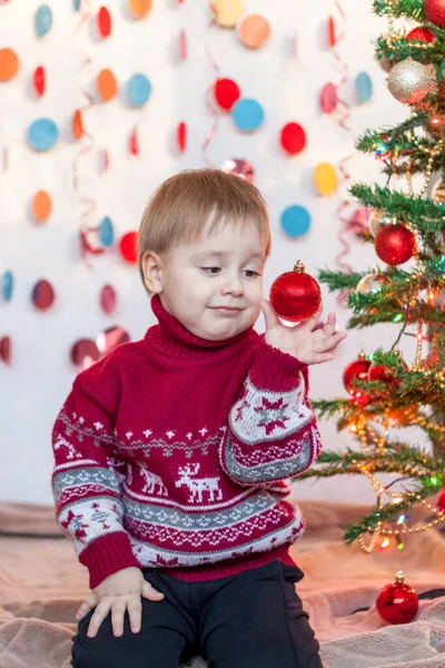 一个小孩坐在一棵优雅的圣诞树下 手里拿着一个圣诞树玩具 孩子们的感情圣诞节和新年 广告横幅的概念 — 图库照片