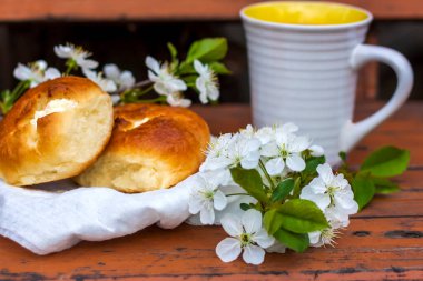 Koyu renkli, yıpranmış ahşap bir masada fırınlanmış ekmek ve bir fincan kahve. Kompozisyon beyaz çiçeklerle süslenmiş bir dal ile süslenmiş. Kiraz ağacı çiçekleri. Seçici odaklanma. Bulanık arkaplan.