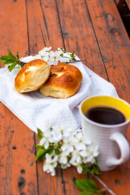 Koyu renkli, yıpranmış ahşap bir masada fırınlanmış ekmek ve bir fincan kahve. Kompozisyon beyaz çiçeklerle süslenmiş bir dal ile süslenmiş. Kiraz ağacı çiçekleri. Seçici odaklanma. Bulanık arkaplan.