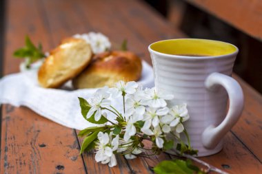 Koyu renkli, yıpranmış ahşap bir masada fırınlanmış ekmek ve bir fincan kahve. Kompozisyon beyaz çiçeklerle süslenmiş bir dal ile süslenmiş. Kiraz ağacı çiçekleri. Seçici odaklanma. Bulanık arkaplan.