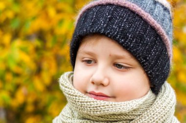 Sonbahar havası. Sarı yapraklardan oluşan bir arka plana karşı duran bir çocuk. Örgü şapkalı bir çocuğun sonbahar portresi. Görüş. Sevimli gülümseyen çocuk. Sonbahar.