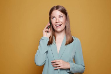 Smiling Caucasian woman looks sideways, has telephone conversation, holds mobile phone near ear, dressed in formal green clothing, discusses last pleasant news, stands iisolated on yellow wall.