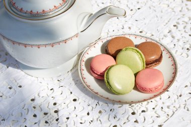 Seramik çay tabağı ve çaydanlık ile makarnalı bisküviler, beyaz işlemeli masa örtüsü.