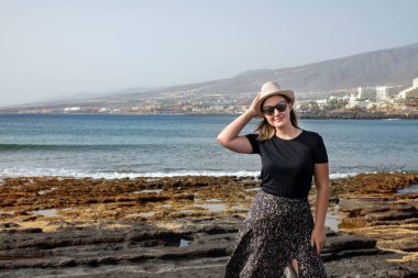 Playa de Las Americas 'ta tek eliyle saman şapkasını tutarken gülümseyen ve kameraya bakan genç bir kadın, tropikal tatil ya da tek başına seyahat kavramı, Tenerife, Kanarya Adaları, İspanya