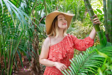 Yazın kırmızı elbise giyen genç ve çekici bir kadının hayali portresi, gözlerini kapalı tutarken geniş kenarlı büyük bir hasır şapka ve huzurlu tropikal yemyeşil bahçenin tadını çıkarıyor.