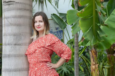 Güçlü bir palmiye ağacına yaslanırken ve yüzünde bir gülümsemeyle kameraya bakarken, bereketli çiçeklerle tropikal bir bahçede dinlenirken, yaz kırmızısı bir elbise giyen genç çekici bir kadın.