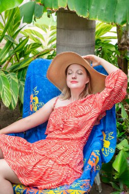 Güzel kırmızı bir elbise giyen, lüks bir bahçede şezlongda oturan, gözleri kapalı ve tek eliyle büyük boy hasır şapkasını tutan çekici beyaz bir kadın.