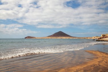 El Medano tatil köyünde bulutlu bir sabah, sakin denize doğru sakin dalgalar ideal yüzme koşullarını ve Tenerife, Kanarya Adaları, İspanya 'da Montana Roja olarak bilinen volkanik dağı gösteriyor.