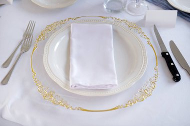 Zarif resmi masa düzeninde cam bir tabak ve süslenmiş altın detaylar üst üste düzgünce katlanmış bir peçeteyle beyaz bir masa örtüsüne yerleştirilmiş ve yanında klasik gümüş çatal bıçak takımı var.