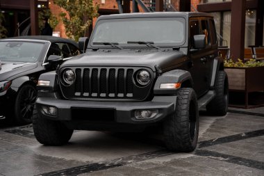 A black Jeep SUV is prominently parked in an urban area, showcasing its rugged design and wide tires. The scene captures the essence of adventure and city life