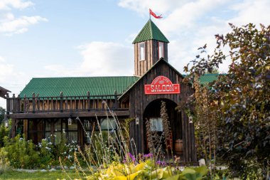 Rustic saloon with wooden structure and green roof, showcasing a classic Western architectural style. Surrounded by lush greenery and colorful flowers under a clear blue sky