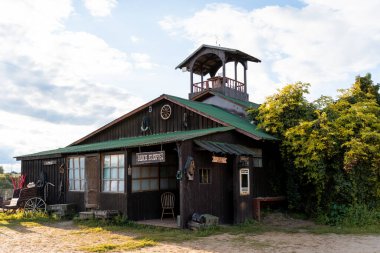Rustic Western ranch outpost featuring a wooden facade and roof. Surrounded by lush greenery, the building exudes an old-fashioned charm, complete with wagon and vintage signs