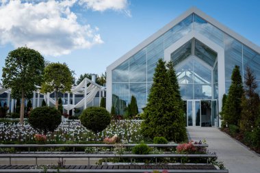 Modern glass-walled greenhouse surrounded by a lush, meticulously landscaped garden with a variety of trees and plants. Bright blue sky and clouds add to the serene atmosphere