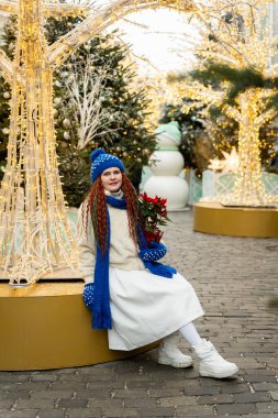 Şenlikli süslemeler ve aydınlatılmış ağaçların arasında otururken, kışın rahat giysiler içinde gülümseyen bir kadın. Bir buket tutuyor, her mevsim neşeyle çevrili, hareketli bir tatil sahnesinde.