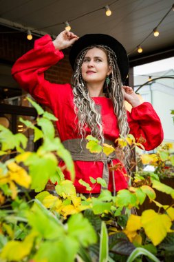 Canlı kırmızı elbiseli bir kadın gür sonbahar bitkilerinin arasında dururken şapkasını düzeltir. Yüzü neşeli ve kaygısızdır. Mevsimsel ortama uyum sağlar.