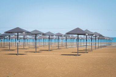 Arka planda mavi deniz bulunan kumlu bir sahildeki boş kumsal şemsiyeleri sakin ve huzurlu bir tatil sahnesi oluşturur. Terkedilmiş atmosfer sezon dışı rahatlamayı öneriyor.