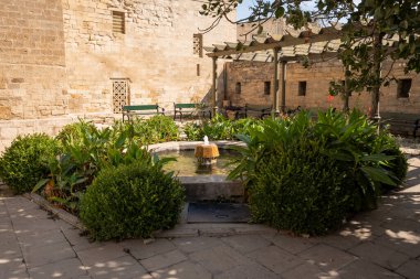 Yeşilliklerle çevrili merkezi bir çeşmenin olduğu taş avlu. Banklar bir pergola altında oturma olanağı sağlar, rahatlamak için mükemmel bir açık hava alanı yaratır.