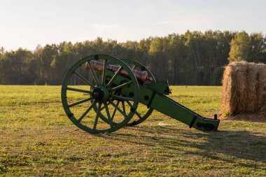 Çimenli bir tarlada, orman zeminli saman balyalarının yanında duran tarihi yeşil top. Huzurlu bir kırsal alan yakalar ve tarih ve sükunet hissi uyandırır.