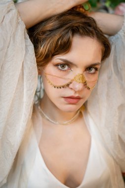 Karmaşık bir şekilde tasarlanmış altın zincirli bir maske takan kadın saçlarını yoğun bir bakışla tutuyor. Görüntü geleneksel mücevherleri vurguluyor ve zarafet ve gizemi çağrıştırıyor.