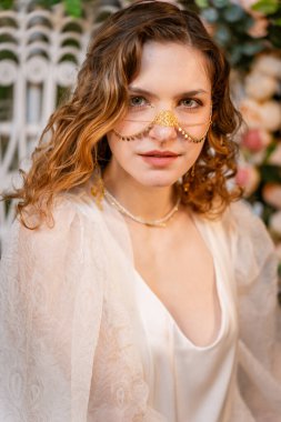 Kadın zarif görünüşünü geliştirmek için altın bir gelin burnu zinciri ile poz veriyor. Beyaz bir bornoz giyer, zarafet ve entelektüellik yayar, çiçekli bir zemine yaslanır.