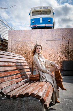 Ahşap bir bankta oturan şık bir ceket ve çizme giyen bir kadın. Arka planda modern bir cephede klasik bir titreşim ile kentsel unsurları harmanlayan klasik bir mavi tren yer alıyor.