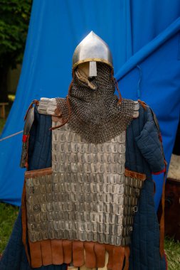 Mavi branda arka planlı metal bir miğfer ve zincir zırhlı ortaçağ zırhı. Zırh birbirine kenetlenmiş metal plakalar, zincir postalar ve deri detaylardan oluşur.