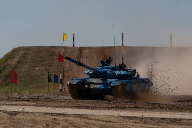Mavi tank toprak yolda hız yapıyor, tozu tekmeliyor ve gücünü sergiliyor. Bayraklar rotayı tank manevraları olarak işaretliyor, hareketin yoğunluğunu yakalıyor.