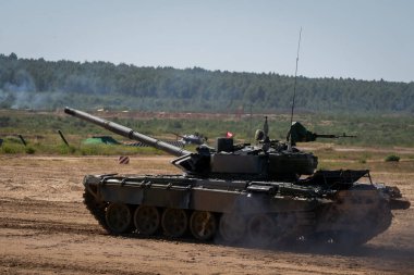 Bir askeri tank tatbikat sırasında tozlu bir alanda ilerler. Güçlü araç engebeli arazide manevra yapıyor, askeri bir ortamda gücünü ve kabiliyetini gösteriyor.