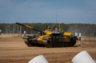 Kamuflaj boyalı bir tank toprak eğitim sahasında yol alıyor, manevra kabiliyeti ve hazırlığını gösteriyor. Orman arkaplanı askeri ortamı ve eylemi ön plana çıkarıyor