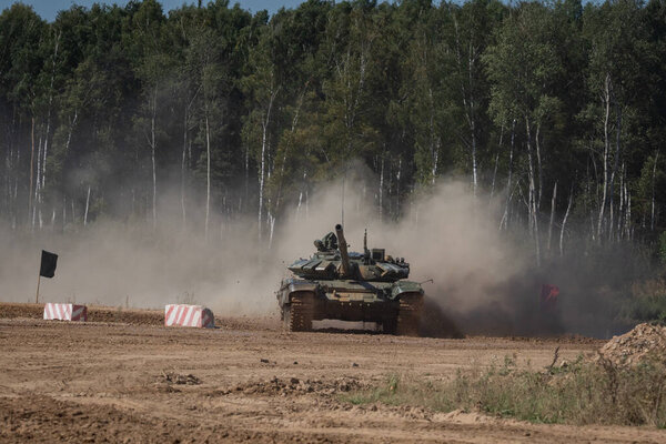 Tank navigating a dusty training field with a forest backdrop. The maneuver creates a cloud of dust as it progresses on the terrain, showcasing military strength and precision