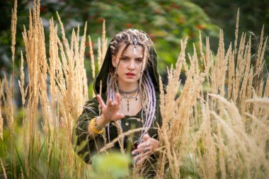 Doğadaki kadın elini uzatarak mistik bir atmosfer yaratıyor. Uzun otlarla çevrili, yeşil bir pelerin giyiyor ve saçlarını boncuklarla örüyor, sihirli bir his veriyor.