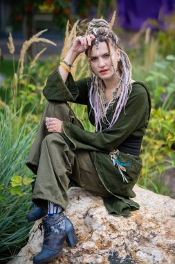 Rasta saçlı genç bir kadın doğal bir ortamda bir kayanın üzerinde oturuyor, günlük giysiler giyiyor ve sakin ve düşünceli bir ifade sergiliyor. Etrafı yeşilliklerle çevrili, bohem bir tarzı benimsiyor.