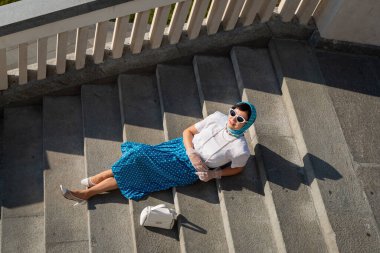 Mavi başörtüsü ve güneş gözlüklü kadın taş merdivenlerde dinleniyor ve güneşin tadını çıkarıyor. Yüzü neşeli, huzurlu ve huzurlu bir anın tadını çıkarıyor.