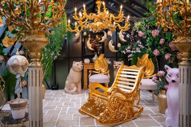 Altın bir avize, karışık sallanan sandalye ve dekoratif heykellerin bulunduğu gösterişli bir oda. Müsrif çiçek düzenlemeleri ve canlı dekorlar zarafet ve ihtişam hissi yaratır.