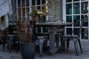 Kırsal ahşap bir masa ve metal sandalyelerin yer aldığı açık hava yemek alanı. Bir tencerede canlı bir sonbahar çiçeği aranjmanıyla süslenmiş. Dekor rahat ve davetkar bir atmosfer yaratıyor.