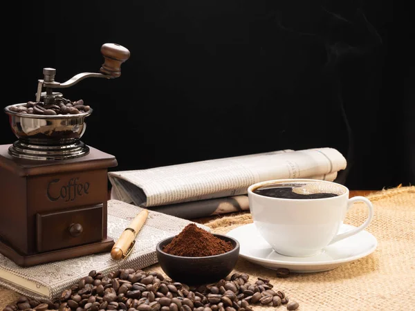 Steam white coffee cup with grinder, roasted beans,coffee ground,newspaper  and note book over burlap hessian  on grunge wood table background