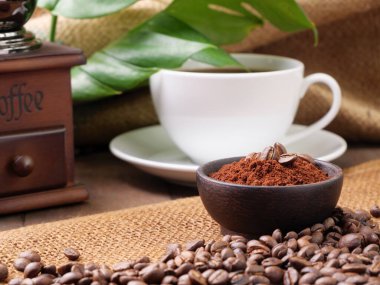 Beyaz kahve fincanları, öğütülmüş kahve, kavrulmuş kahve çekirdekleri, öğütücü ve canavar, çuval bezi arka planda, ahşap masadan ayrılırlar.