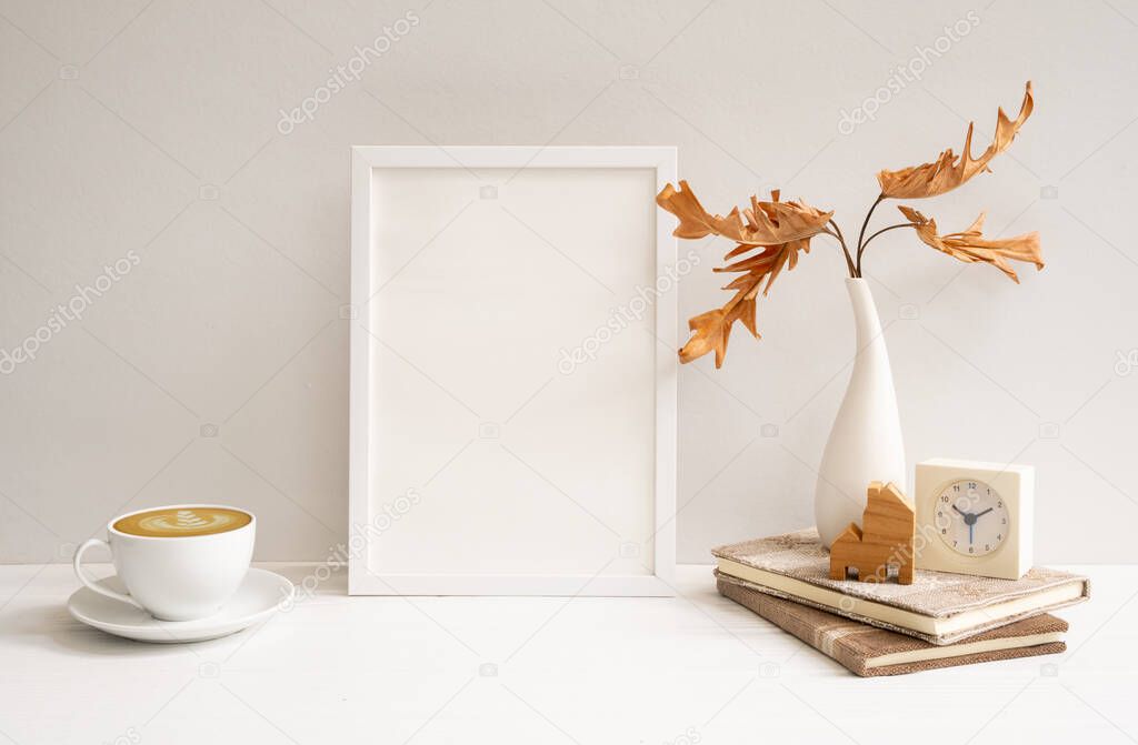 Work space with mock up white wood poster frame, coffee cup,Philodendron dried leaf in vase,clock, books, house model on beige table and wall,modern stylish home decor interior