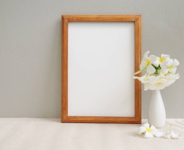 Poster ahşap çerçeve ve güzel Plumeria ya da Frangipani tropikal çiçeklerini bej masa ve çimento duvar arka planında modern beyaz vazoda taklit et. 
